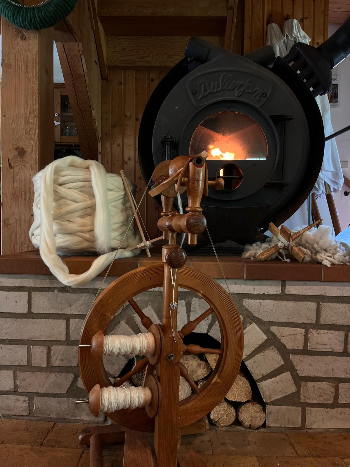 Historisches Spinnrad mit Wolle vor einem brennenden Kachelofen in einer rustikalen Stube.