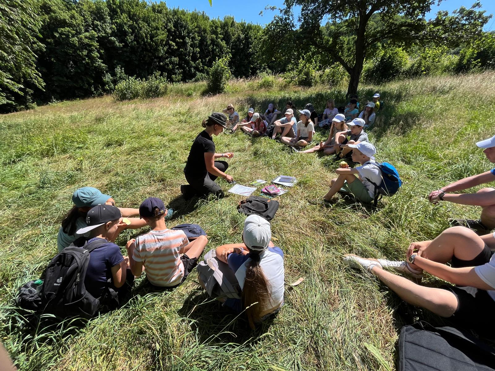 Eine Gruppe von Studierenden sitzt im Kreis auf einer Wiese und folgt einer Unterrichtsstunde im Freien unter Anleitung einer Kursleiterin.